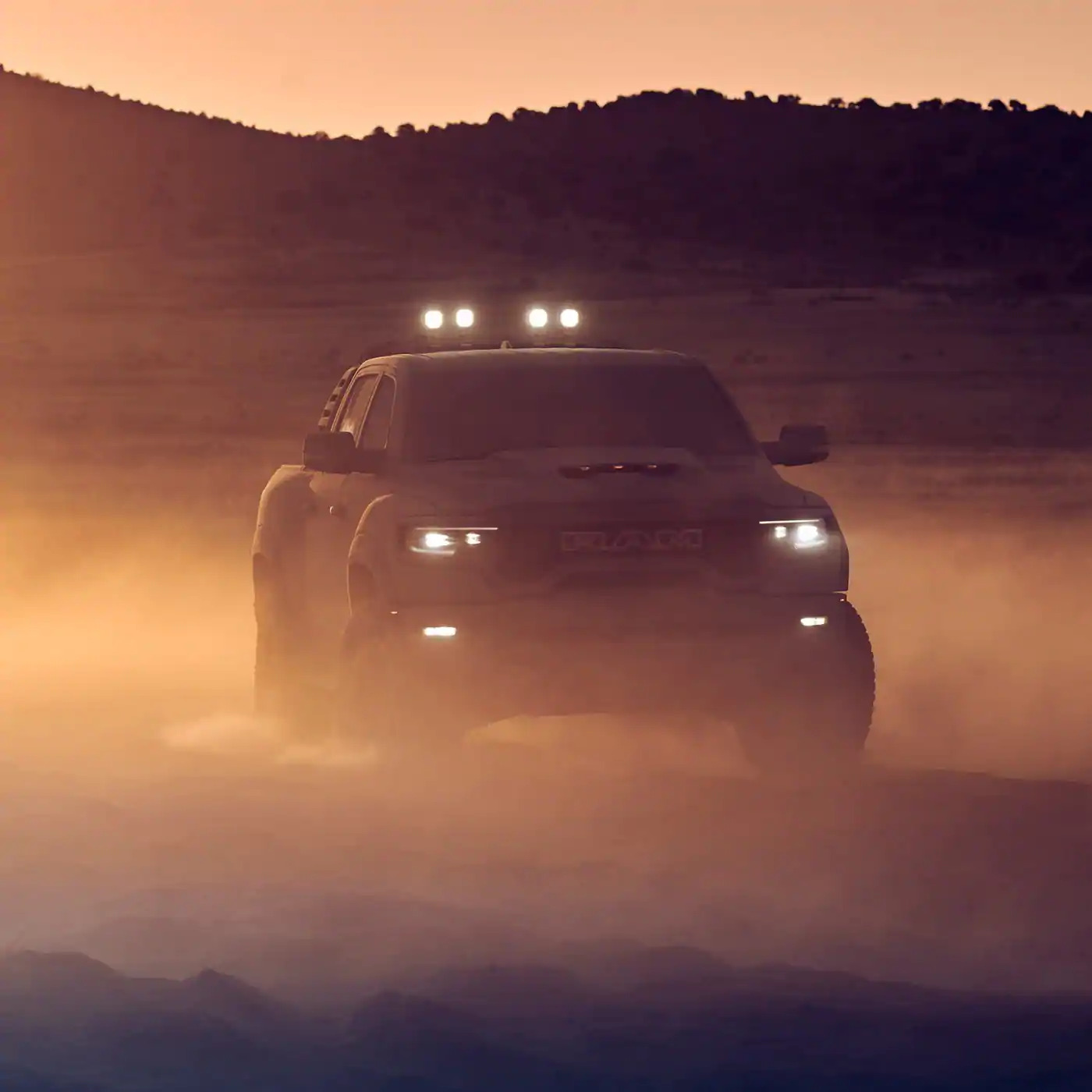 The headlamps of the 2021 Ram 1500 TRX lit as it is driven off-road at sunset.